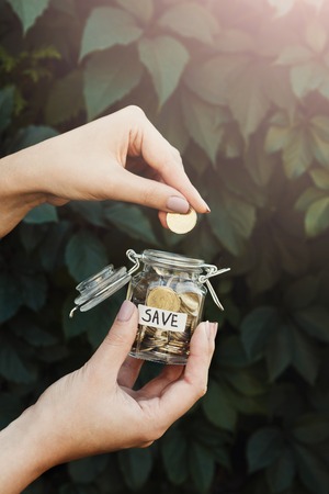 Hand putting coins in glass jar with SAVE label on green leaves background. Money saving for house, dream, vacation. Economic crisis and financial stability conceptの写真素材