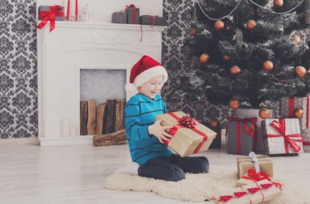 Cute happy boy in santa hat unwrap christmas present box on holiday morning in beautiful room interior. Male child open Xmas gifts near big decorated fir tree and fireplace. Winter holidays conceptの写真素材