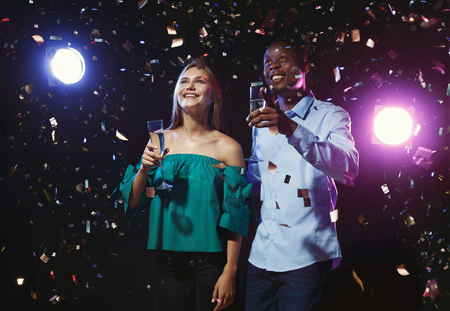 Happy male and female multiethnic friends with champagne flutes at night club background with confetti. Nightlife, birthday or any party conceptの写真素材