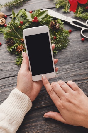 Preparing for christmas background. Female hands holding smartphone with empty screen for copy space against fir tree branches wreath and wooden rustic table backdrop. Holiday online shoppingの写真素材