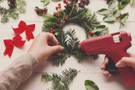 Leisure, tools and trinkets for xmas holiday decoration. Crop of white wooden table background with woman hands with glue gun making craft wreathの写真素材