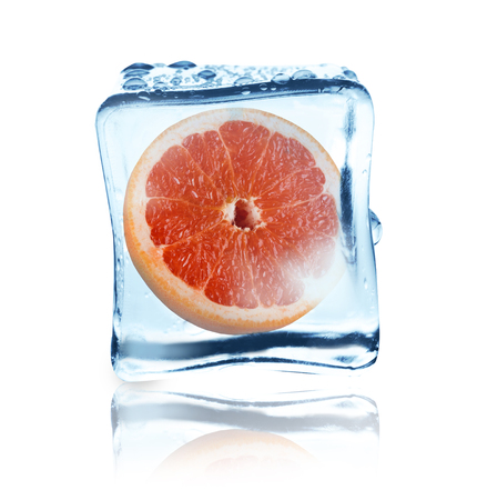 Juicy red grapefruit half frozen inside transparent ice cube, covered with water drops, isolated on white backgroundの写真素材