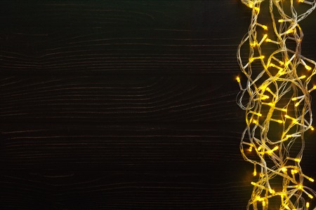 Christmas lights background, copy space. Holiday shiny yellow garland border top view on dark wooden planks surface. Xmas tree decorations, winter holidays illuminationの写真素材