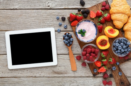 Breakfast background. Morning meals with muesli, croissants, yougurt and berries with wooden cutlery and tablet with empty screen for copy space. Online order of fresh organic food, top viewの写真素材