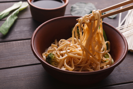 Chopsticks, taking rice noodles with soy sauce in bowl on wooden background, close-up.の写真素材