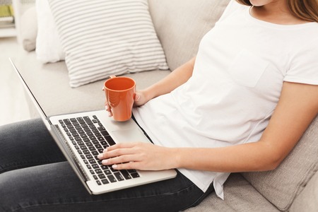 Female hands on typing on laptop. Unrecognizable woman working on computer while sitting on the beige couch at home, cropの写真素材