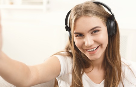 Pretty smiling girl in headphones making selfie, long-haired blonde in white clothes, closeupの写真素材