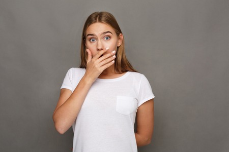 Shocking news. Surprised woman covering mouth with hands while posing to camera on gray studio...