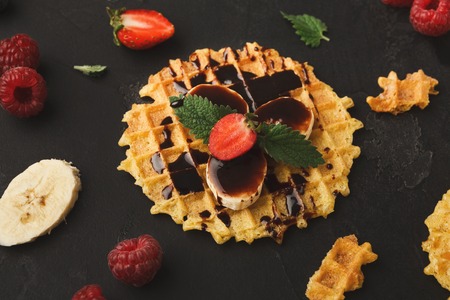 Belgian waffles with fruits and chocolate on black background. Closeup of checkered biscuit with strawberries, banana and topping. Sweet food and tasty breakfast conceptの写真素材