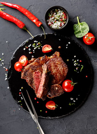 Rare rib eye steak on dark plate decorated with tomatoes, chilli, spices and meat fork, top viewの写真素材