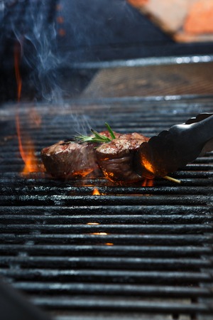 Man cooking meat steaks on professional grill outdoors. Male hand with tongs flipping beefsteaks on...