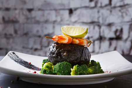 Excuisite restaurant food background. Dorado fillet wrapped in nori on broccoli pillow with shrimps and lemon piece on topnon white platter, sprinkled with pepper salt. Copy space on white brick wallの写真素材