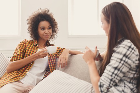 Two happy young female friends with coffee cups talking in living room at home, chatting about their life and relations, gossip and slumber party concept, copy spaceの写真素材