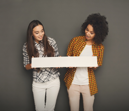 Multiethnic girls holding blank white banner with copy space on gray studio backgroundの写真素材