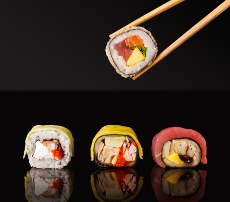 Set of three sushi rolls with fish, vegetables and cheese, chopsticks taking one piece. Traditional japanese food on black glass with reflection, copy space. Asian restaurant meals deliveryの写真素材