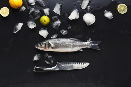 Raw sea bass with ice and knife at black background. Minimalistic mokeup for seafood restaurant or fish market. Top view, copy spaceの写真素材