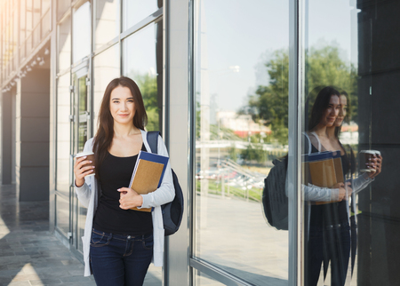 Beautiful brunette student girl with textbooks and coffee walking down at university building, copy spaceの写真素材