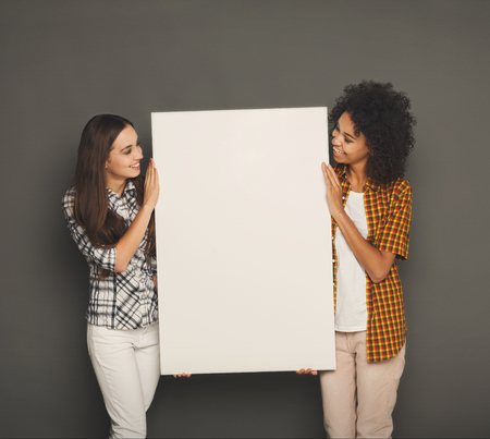 Multiethnic girls holding blank white banner with copy space on gray studio backgroundの写真素材