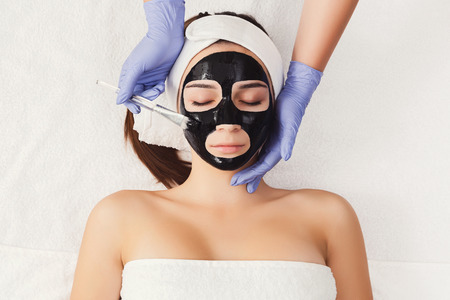 Apply black face mask, spa beauty treatment and skincare. Woman getting facial cleansing mask by...
