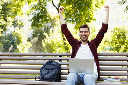 Happy successful man with arms raised in air. Young casual student with laptop celebrating victory outdoors, sitting on bench in park. Winner and achievement concept.の写真素材