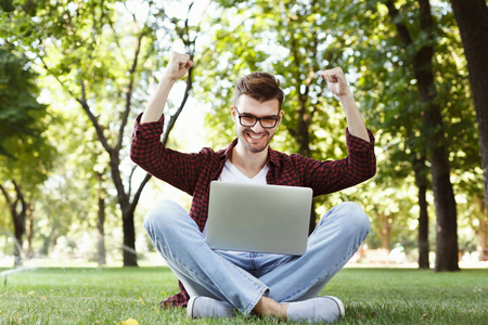 Happy successful man with arms raised in air. Young casual student with laptop celebrating victory outdoors, sitting on grass in park. Winner and achievement concept.の写真素材