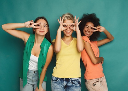 Happy female friends in colourful clothes having fun at blue background. Three young women posing, showing different signs and laughing, slumber party, copy spaceの写真素材