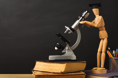 Educational background. Wooden marionette in graduation hat looking in microscope against empty classroom blackboard for copy space. Back to school conceptの写真素材