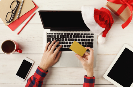 Man shopping online with credit card and laptop with blank screen. Top view of human hands, laptop keyboard, coffee, Santa hat, present on white wooden table background. New Year conceptの写真素材