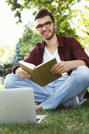 Young attractive student reading book and working on laptop, preparing for exams at university or college outdoors. Education and remote working concept, copy spaceの写真素材