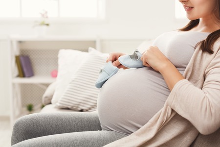 Pregnant woman holding tiny shoes near belly. Smiling expectant woman preparing for baby birth,...