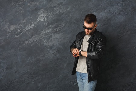 Handsome stylish smiling man in sunglasses and brutal black leather jacket watching time and posing in studio. Fashion boy at grey background, copy spaceの写真素材