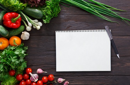 Notepad for ingredients list and border with fresh vegetables on black background. Mockup for healthy dishes recipes, top view, copy spaceの写真素材