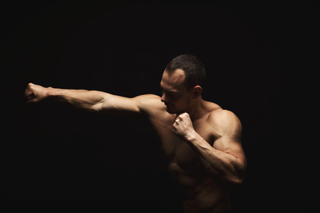 Athletic man make punch. Handsome fitness model show naked torso, muscular body. Strong hands, shoulder muscles and biceps. Studio shot on black background, low key. Kickboxing and fight sport conceptの写真素材