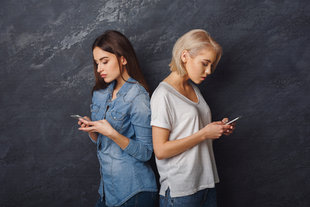 Two casual girls with smartphones at dark studio background. Concentrated women serfing the net or playing online games on mobiles. Gadgets addiction concept, copy spaceの写真素材