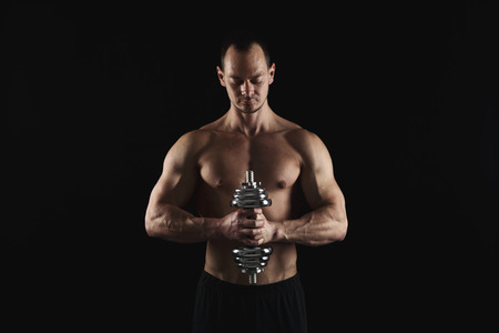 Athletic young man with a dumbbell on a black background. Naked torso, muscular body. Strong chest and shoulder muscles. Studio shot, low key. Bodybuilding concept, copy spaceの写真素材