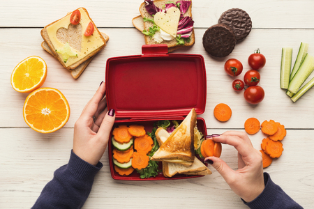 Preparing healthy vegetarian snacks on white rustic wood. Female hands making sandwiches and putting into take away plastic lunch box, top view. Eating right, picnic and food storage conceptの写真素材