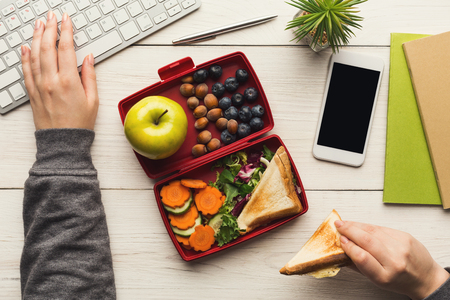 Healthy snack at office workplace. Businesswoman eating organic vegan meals from take away lunch box at wooden working table with computer keyboard and smartphone with empty screen for copy spaceの写真素材