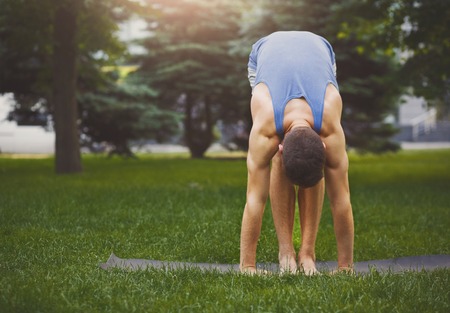 Sporty man doing Standing Forward Bend posture outdoors. Guy practicing yoga, stand head to knees in park, copy spaceの写真素材