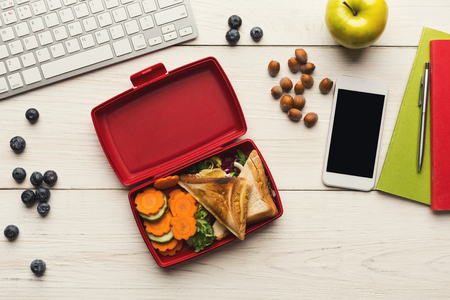 Healthy snack at office workplace. Eating organic vegan meals from take away lunch box at wooden working table with computer keyboard and smartphone with empty screen for copy space, top viewの写真素材