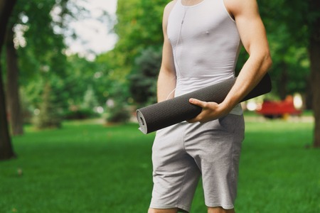 Unrecognizable athlete holding mat and listening to music in park, copy space. Serene sporty guy preparing for workout outdoorsの写真素材