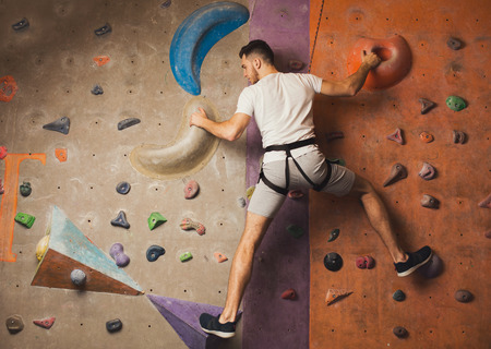 Young happy man practicing rock-climbing on artificial wall indoors. Active lifestyle and bouldering...