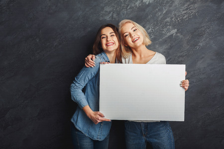 Two girlfriends holding blank white banner at dark studio background. Young women showing empty board with copy space for advertisementの写真素材