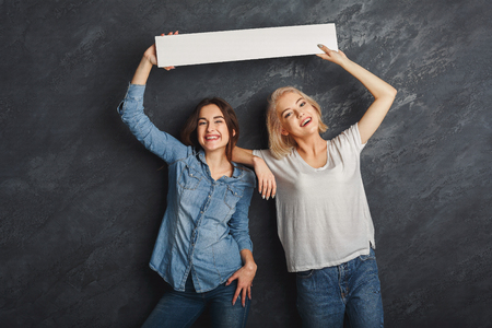 Two girlfriends holding blank white banner over heads at dark studio background. Young women showing empty board with copy space for advertisementの写真素材