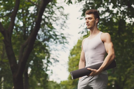 Young athlete holding mat and listening to music in park, copy space. Serene sporty guy preparing for workout outdoorsの写真素材