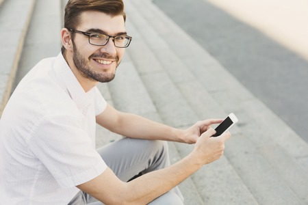 Smiling businessman messaging on smartphone outdoors. Young happy salesman working with mobile sitting on stairs in urban area, copy spaceの写真素材