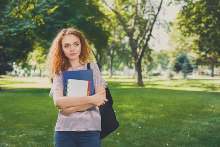 Young redhead woman outdoors. Female student standing in the park with an exercise book and backpack. Education conceptの写真素材