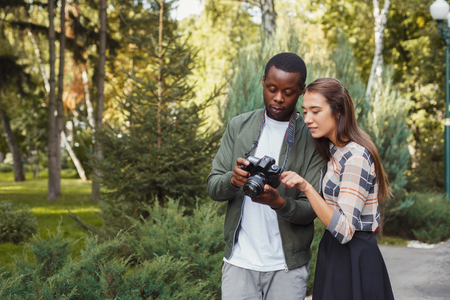 Multiethnic couple watching pictures on camera, standing in park, copy spaceの写真素材