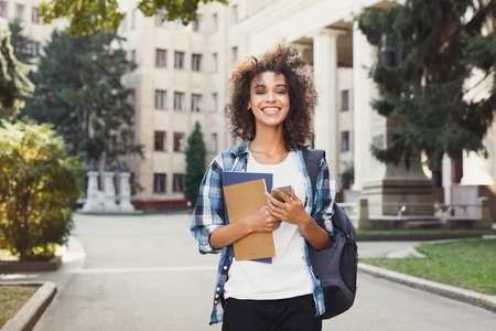 Happy smiling african-american student girl with backpack at university background. Woman with mobile and workbooks having rest in campus during lunch break. Technology, education, leisure conceptの写真素材