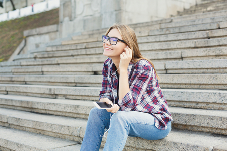 Young smiling woman sitting on university stairs, listening to music with earphones outdoors. Rest, education and relax concept, copy spaceの写真素材