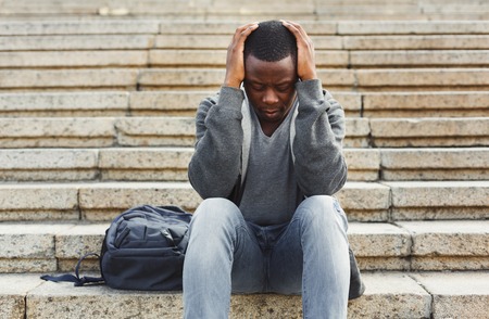 Desperate african-american student sitting on stairs outdoors in university campus. Man raised hands to his head. Migraine, education and overworking concept, copy spaceの写真素材